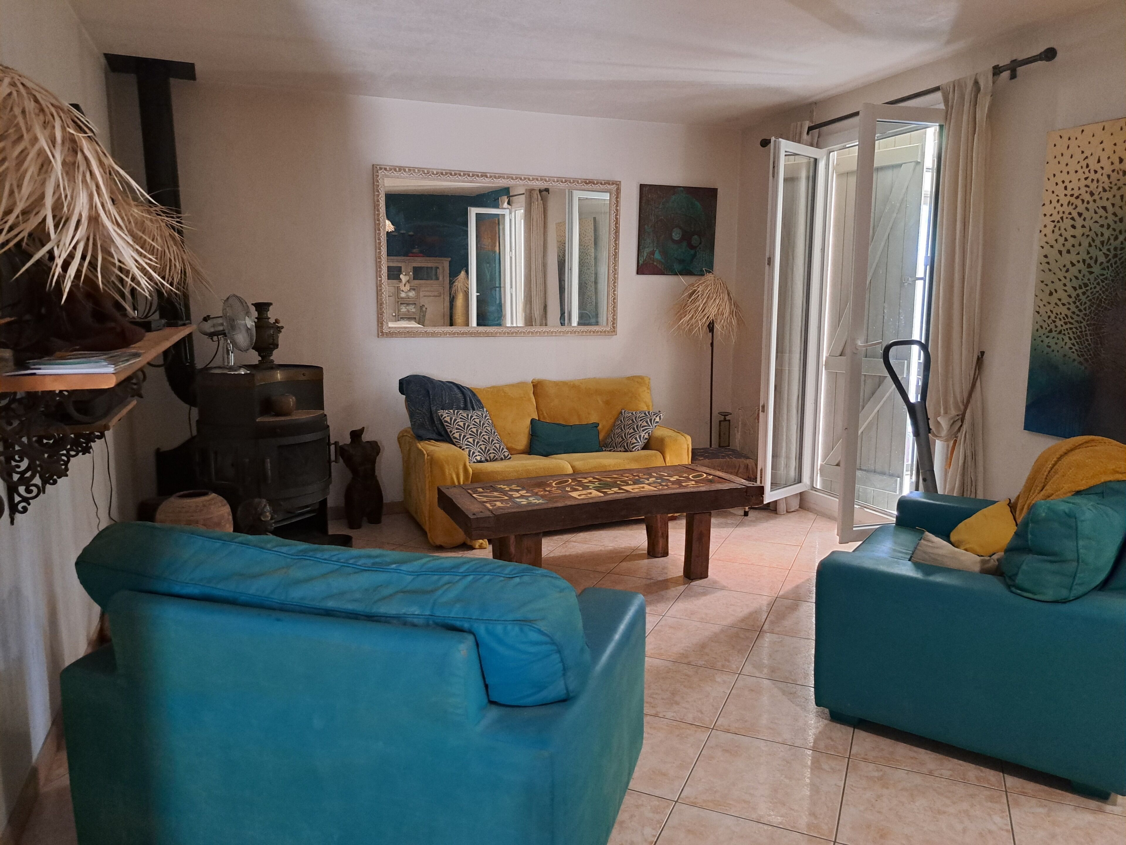 A living room with a yellow sofa, blue armchairs, a wooden coffee table, a fireplace, a mirror, and a large window with curtains.