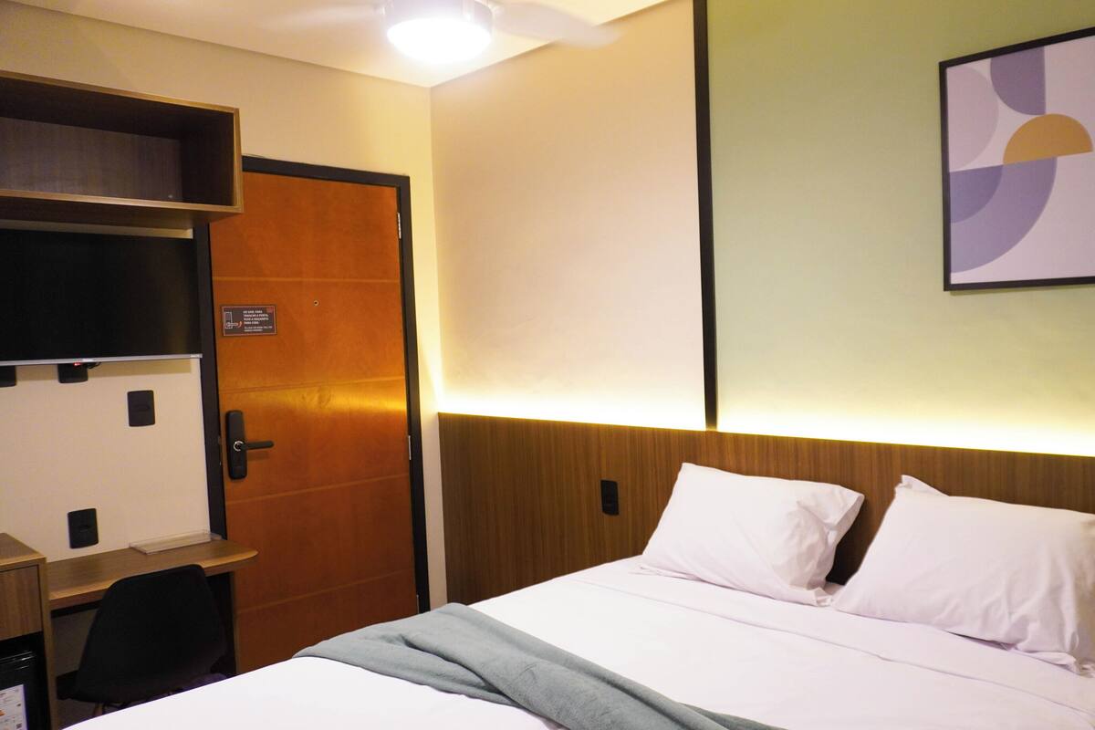 A hotel room with a bed, a desk, a chair, a wooden door, and a wall-mounted artwork.