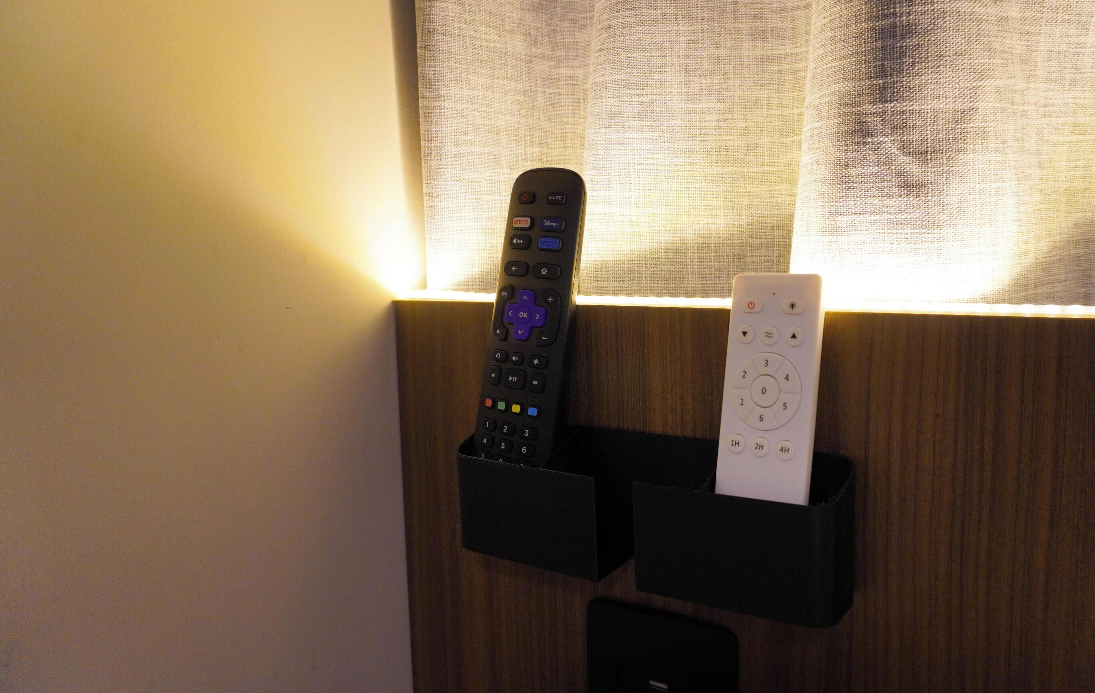Two remote controls in a black holder, a window with sheer curtains, and a wooden wall.