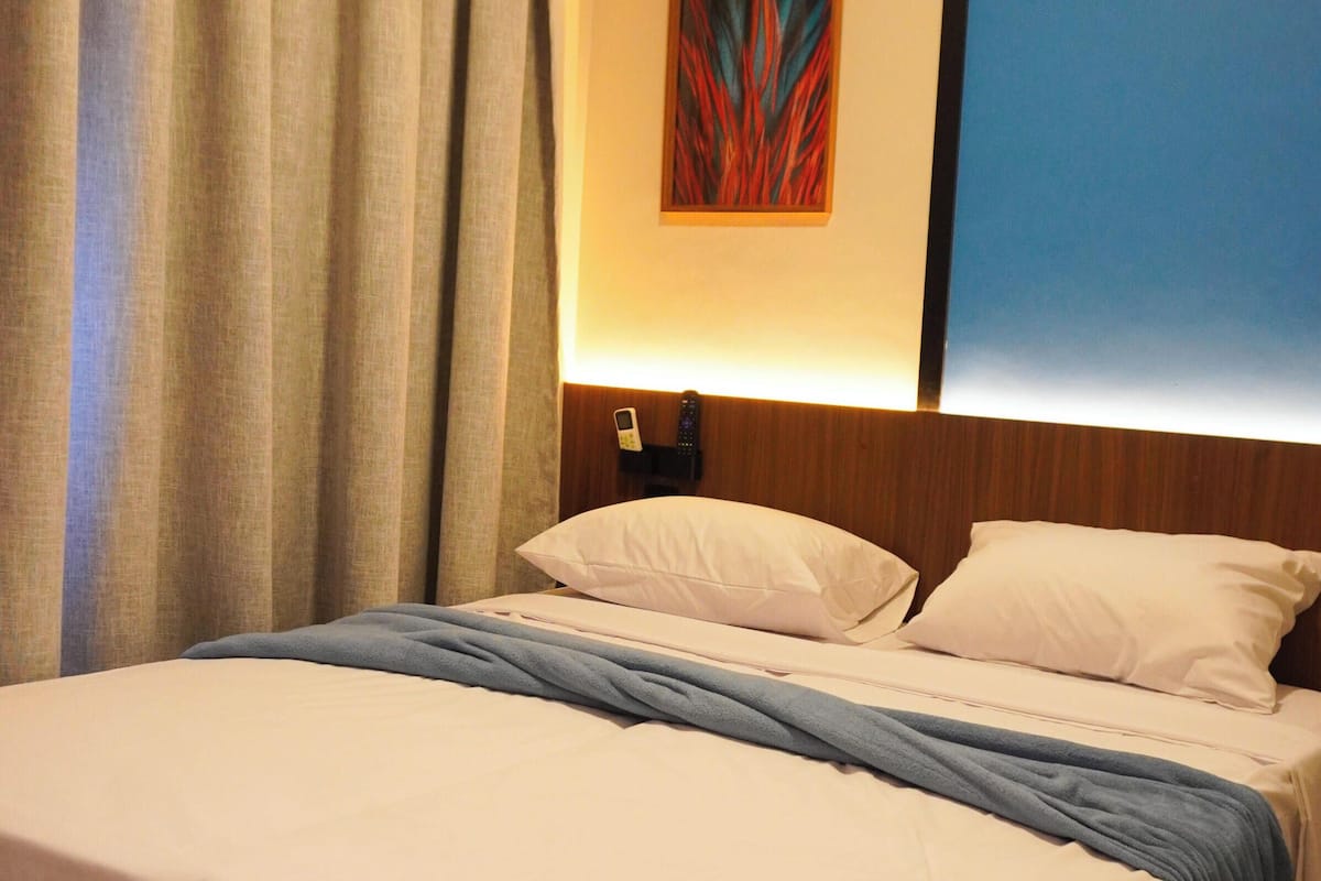 A hotel room with a bed, curtains, a painting, and a bedside table with a phone.