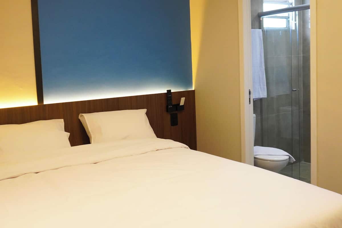 A hotel room with a bed, a nightstand, a wall-mounted remote control, and a shower area.