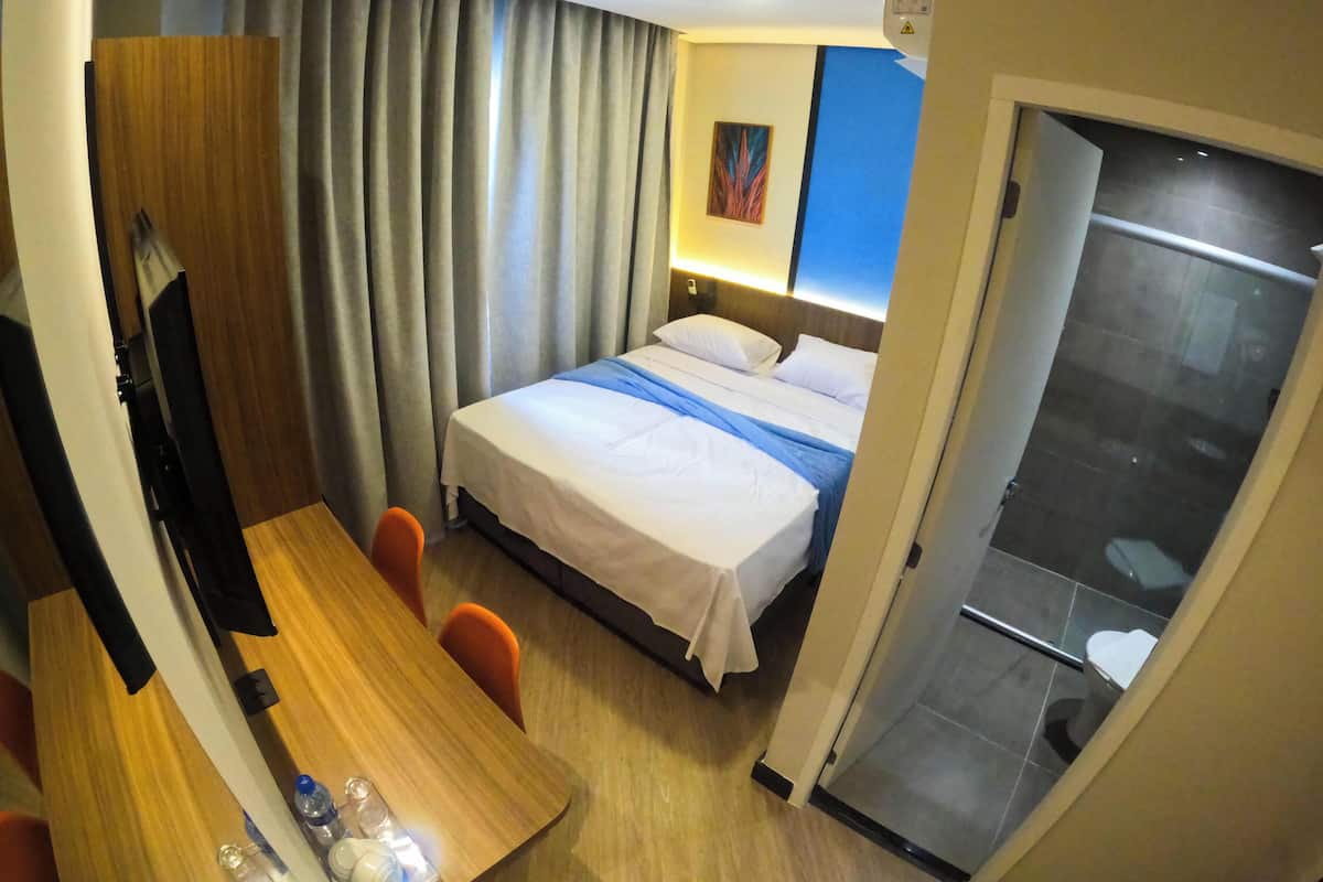 A hotel room with a bed, a desk, a chair, a shower, and a painting on the wall.