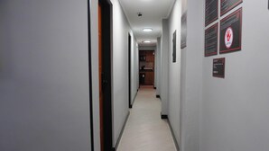 Un pasillo con piso de color claro, paredes blancas y una puerta a la izquierda. Hay letreros en la pared derecha, incluyendo uno con un símbolo de mano y texto.