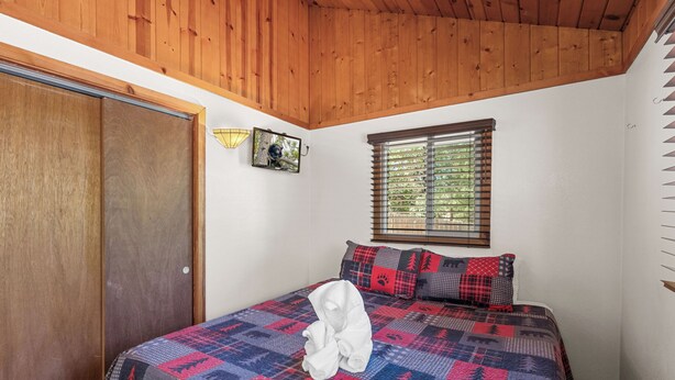 Een slaapkamer met een houten plafond, een bed met quilt, een raam met zonwering en een wandlamp.