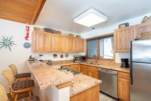 A kitchen with wooden cabinets, granite countertops, a stainless steel refrigerator, and a window with a view. - Riverside Condos B101 2 Bedroom Condo (Telluride)