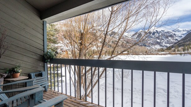 Una terraza de madera con sillas y una mesa, con vista a un paisaje nevado y montañas.