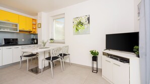 Eine moderne Küche mit weißen Schränken, einem Esstisch mit Stühlen, einem schwarzen Fernseher und einem Fenster mit Jalousien.