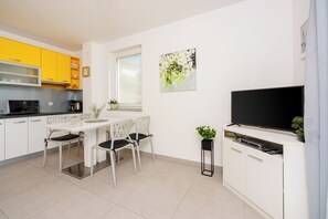 A modern kitchen with white cabinets, a dining table with chairs, a black TV, and a window with blinds. - Vacation apartment for 2-5 people with sea view (Dramalj)