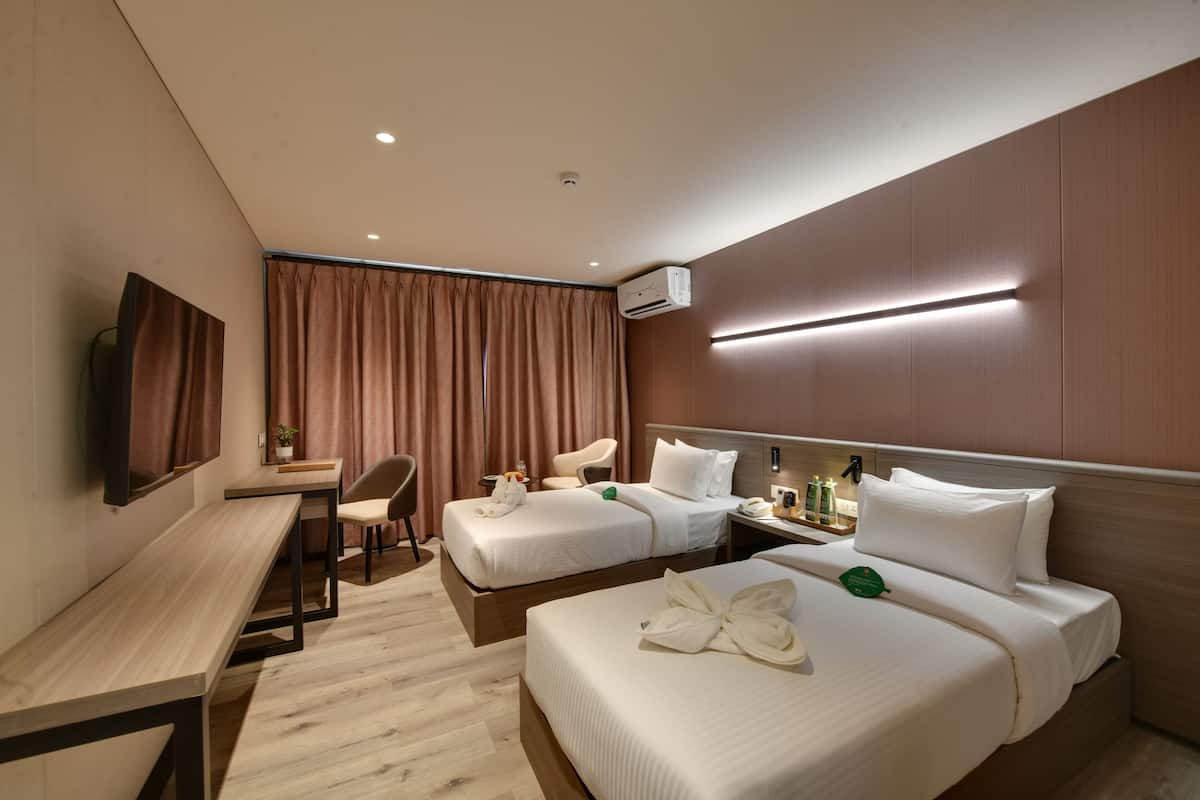 A hotel room with two beds, a desk, a chair, a TV, and an air conditioning unit.