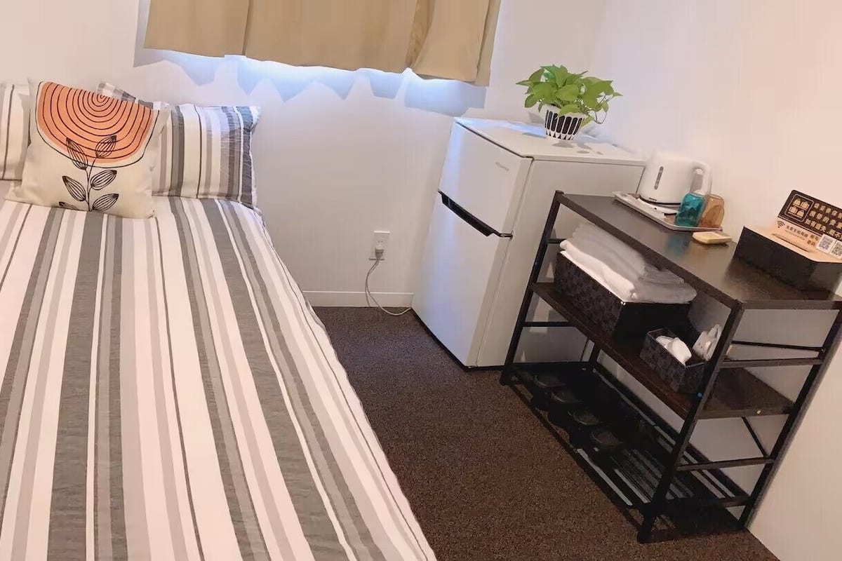 A small room with a bed, a desk, a refrigerator, and a plant.
