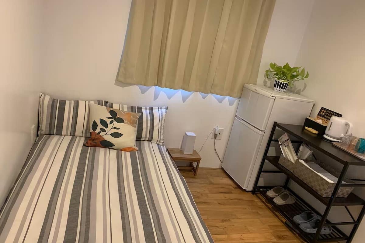 A small, single-room accommodation with a bed, a small table, a refrigerator, a shelf with shoes, and a potted plant.