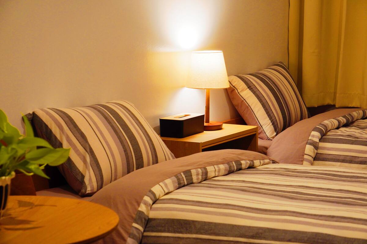 A hotel room with a bed, a bedside table with a lamp, and a small table with a plant.