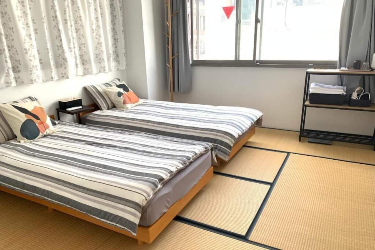 A room with two beds, a window with curtains, a shelf with items, and tatami flooring.