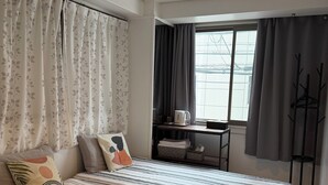Ein Schlafzimmer mit Bett, Kissen, einem Fenster mit Vorhängen, einem kleinen Tisch mit Wasserkocher und einer Stehlampe.