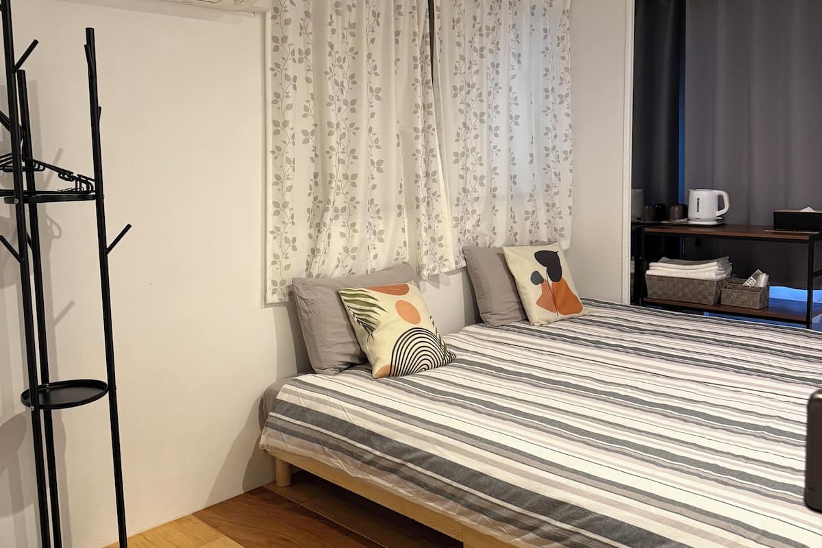 A bedroom with a bed, a black coat rack, a window with curtains, and a bedside table with a kettle.