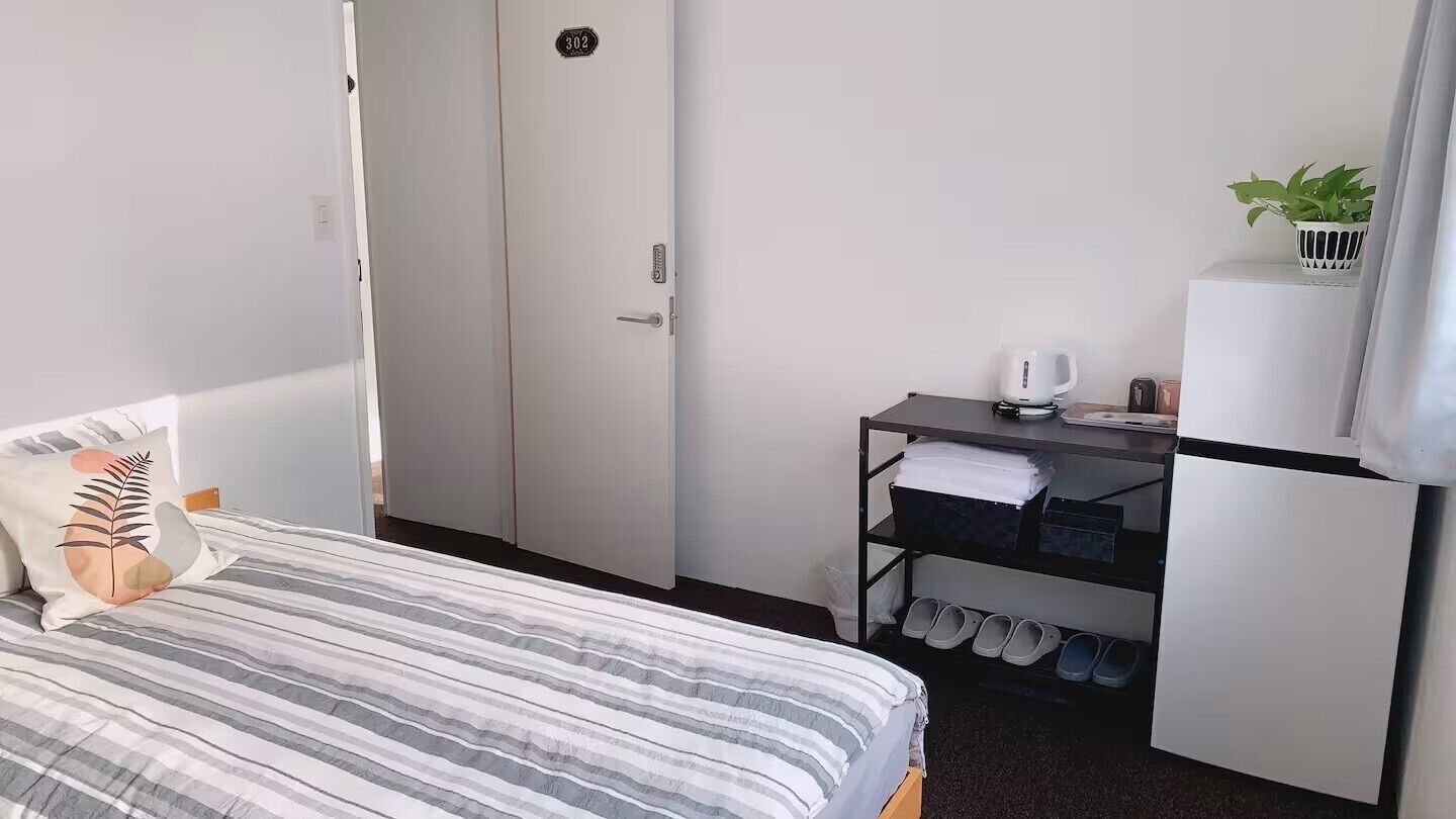 A hotel room with a bed, a desk with a coffee machine, a shoe rack, and a small plant.