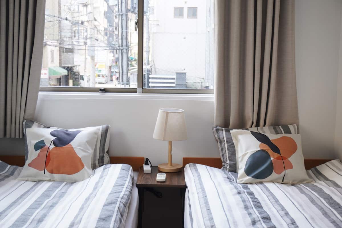 A hotel room with two beds, a nightstand, a lamp, and a window with a city view.