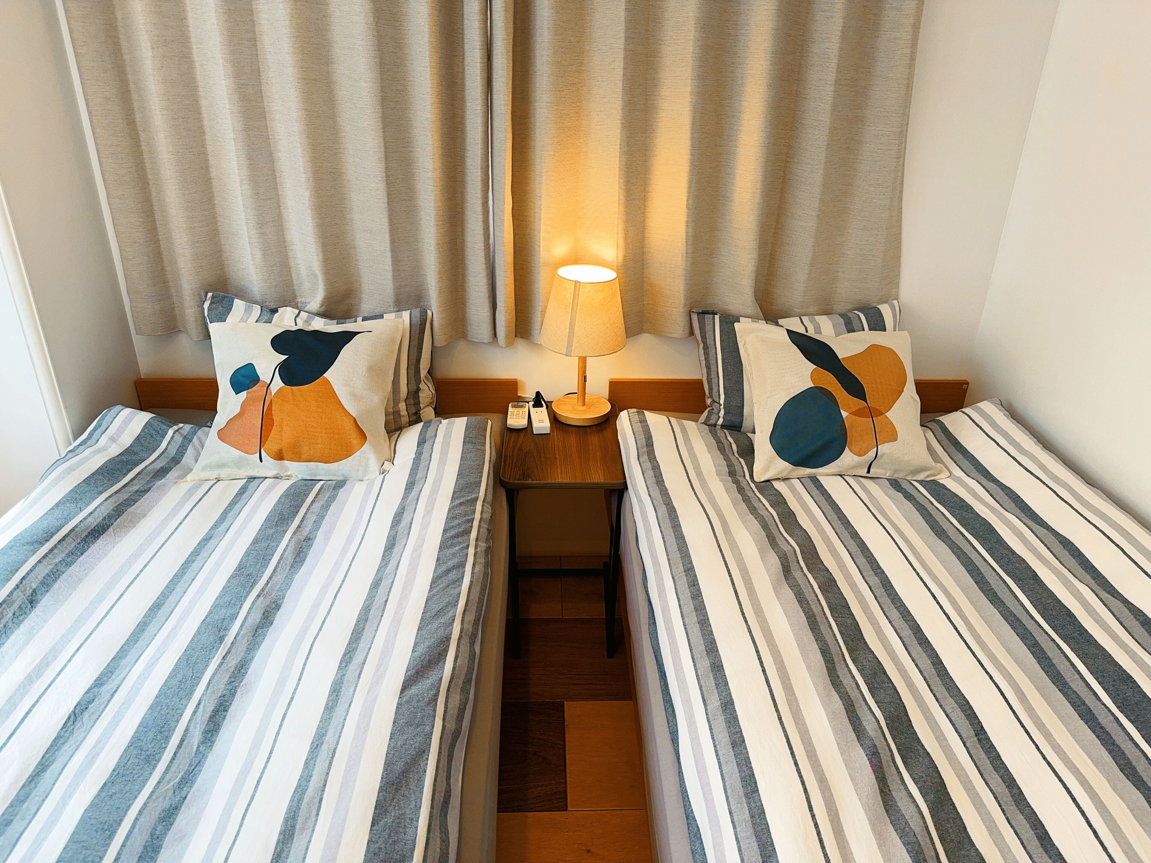 A room with two single beds, a bedside table with a lamp, and a window with curtains.