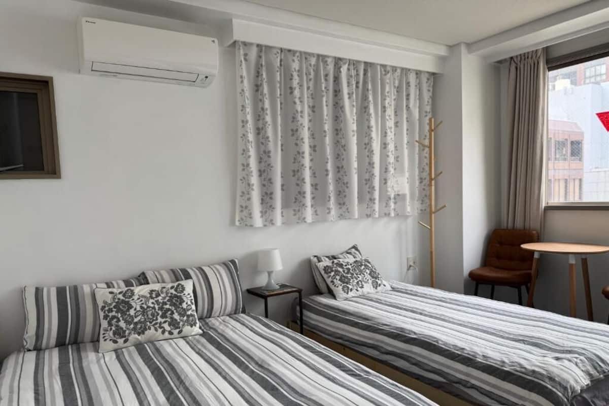 A hotel room with two beds, a window with curtains, a chair, a table, and an air conditioning unit.