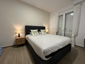 Ein Schlafzimmer mit einem großen Bett, zwei Nachttischlampen, einem Holznachttisch und einem Fenster mit Vorhängen.