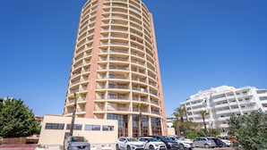 A tall, multi-story hotel with a curved facade, a parking lot with several cars, and a clear blue sky. - Apartment 'Praia Azul Ocean View' with Sea View, Wi-Fi and Air Conditioning (Portimão)