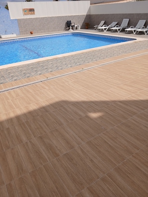 Ein Pool mit gefliestem Rand, ein sonnendurchfluteter Deck mit Ablassgitter und Liegestühle entlang des Beckens.
