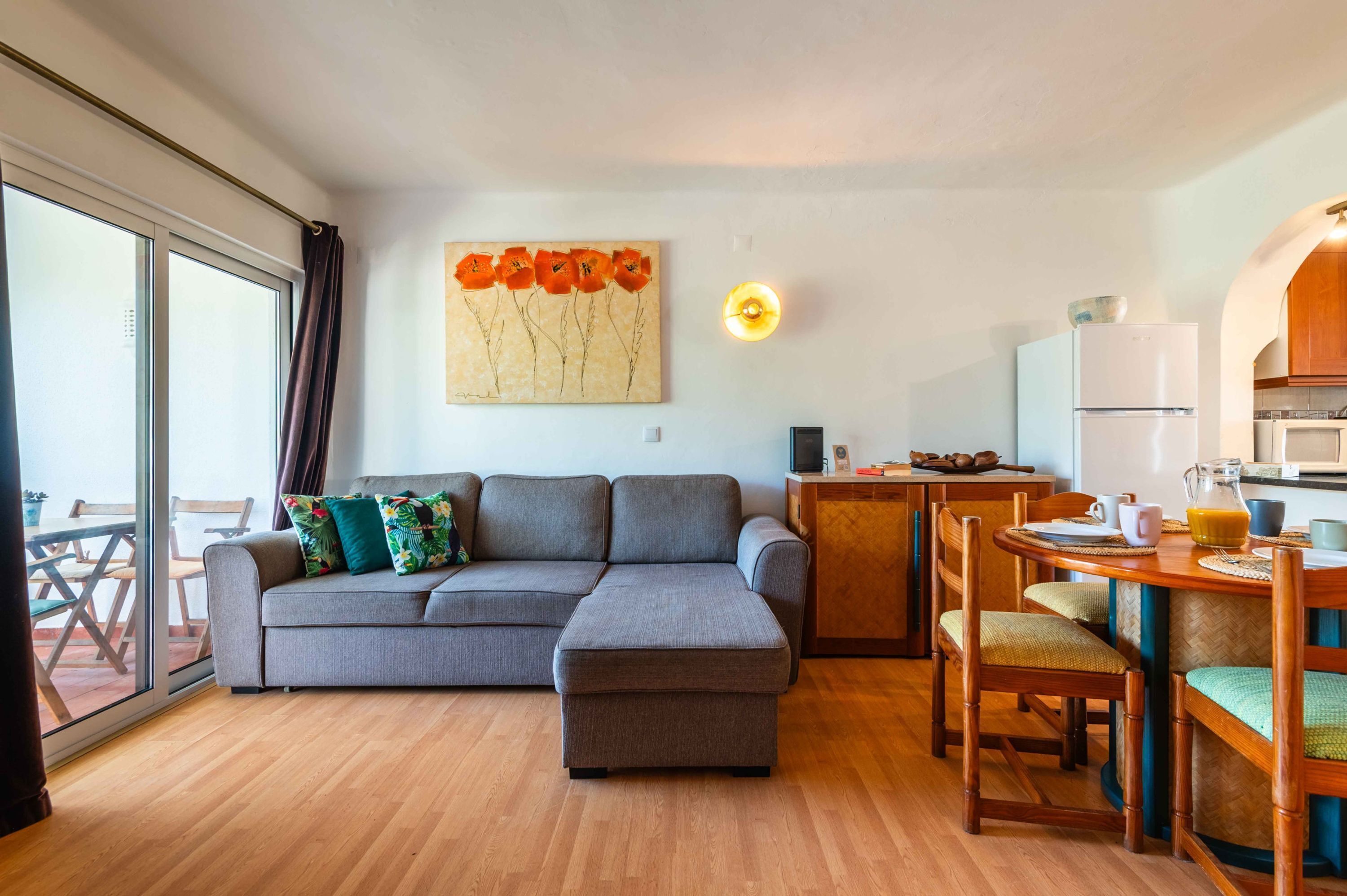 Ein modernes Wohnzimmer mit einer grauen Eckcouch, einem Esstisch mit Stühlen, einer Küchenzeile inklusive Kühlschrank und einem großen Fenster mit Vorhängen.