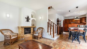 Ein geräumiges Wohnzimmer mit Kamin, Korbmöbeln, einem Holzesstisch und einer Küchenzeile mit Holzausbau und Ziegelrückwand.