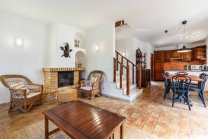 A spacious living room with a fireplace, wicker furniture, a wooden dining table, and a kitchen area with wooden cabinets and a brick backsplash. - Casa das Conchas V4 Praia da Coelha (Albufeira)