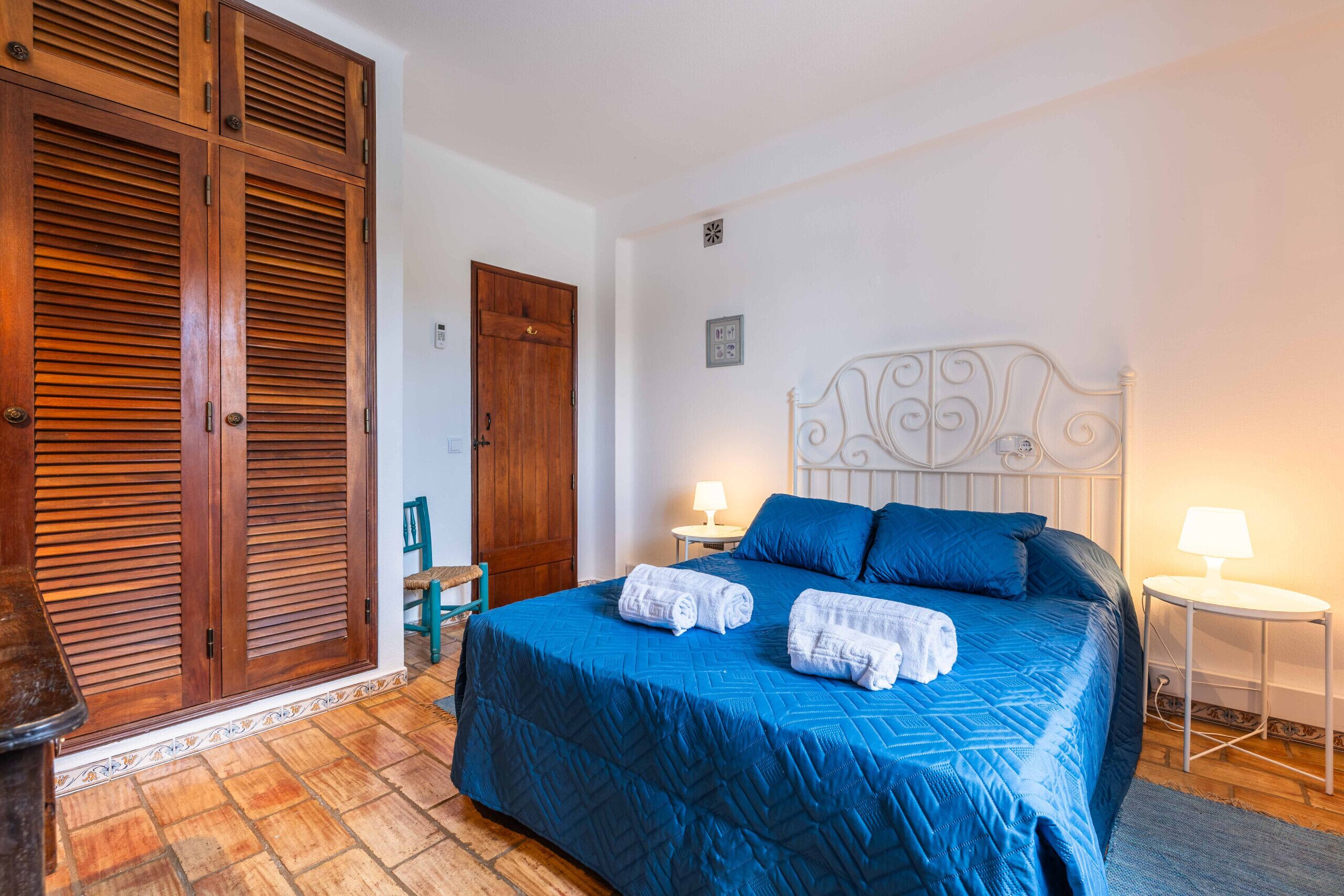 A bedroom with a large bed, a wooden wardrobe, a white metal headboard, and two bedside tables with lamps.