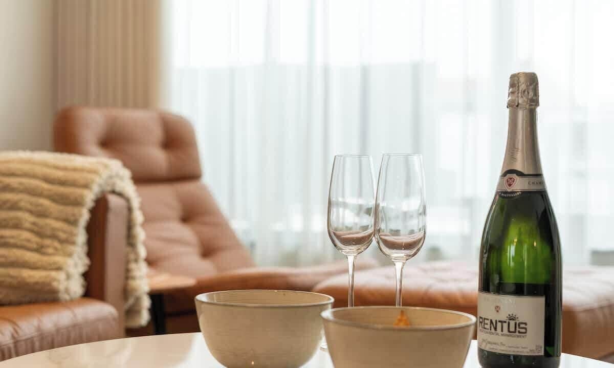 En flaska RENTUS-champagne, två glas och två skålar står på ett bord i ett rum med en fåtölj klädd i läder och ett fönster med gardiner.