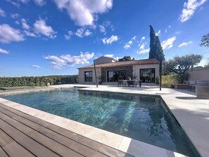 A swimming pool with a wooden deck, a stone patio, and a building with a tiled roof and large windows. - Stunning modern villa with views of Sainte Victoire, nestled between the sea and the mountains (Cabriès)