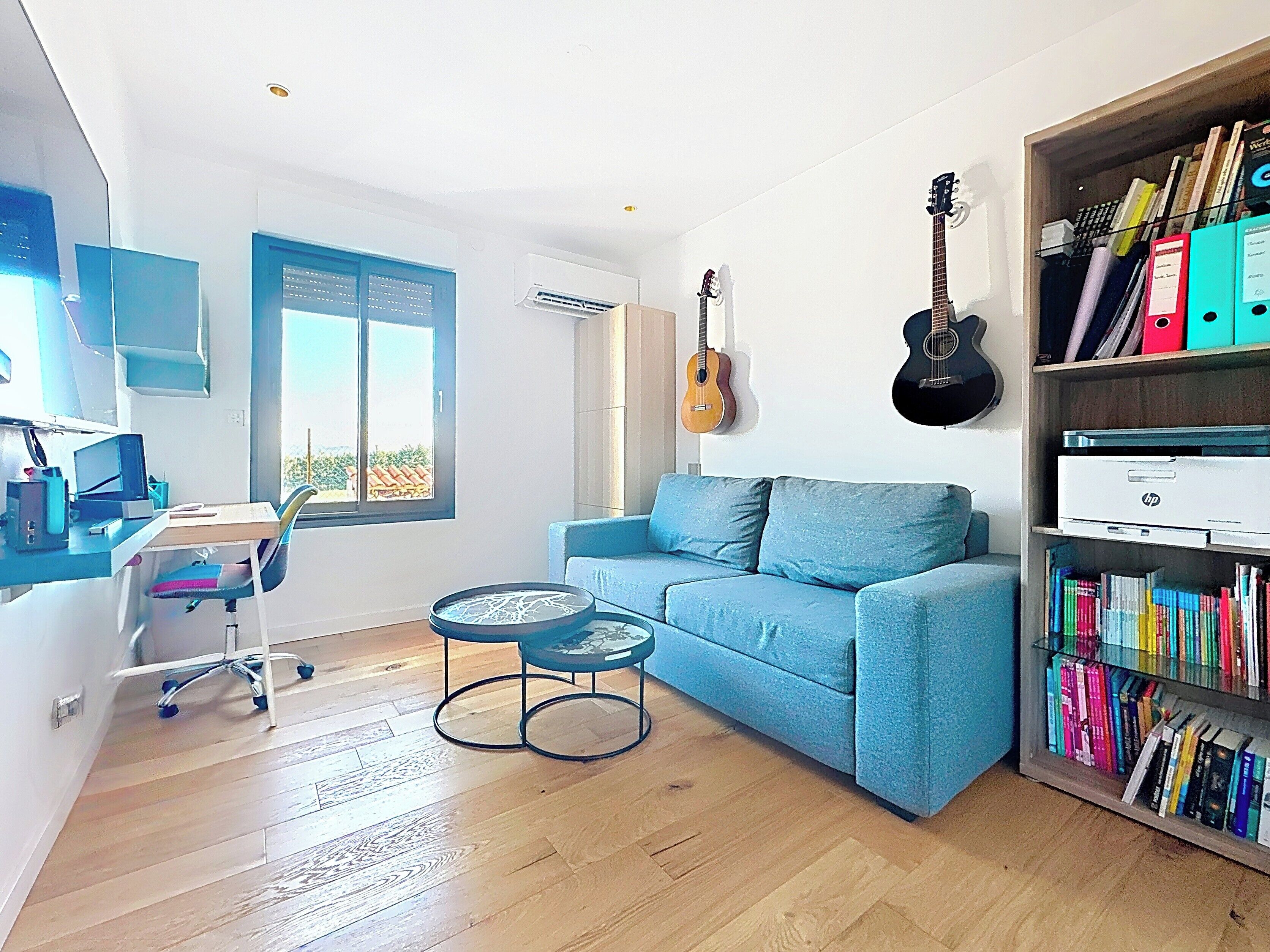 A room with a blue sofa, a small round table, a bookshelf, a guitar, and a desk with a computer.