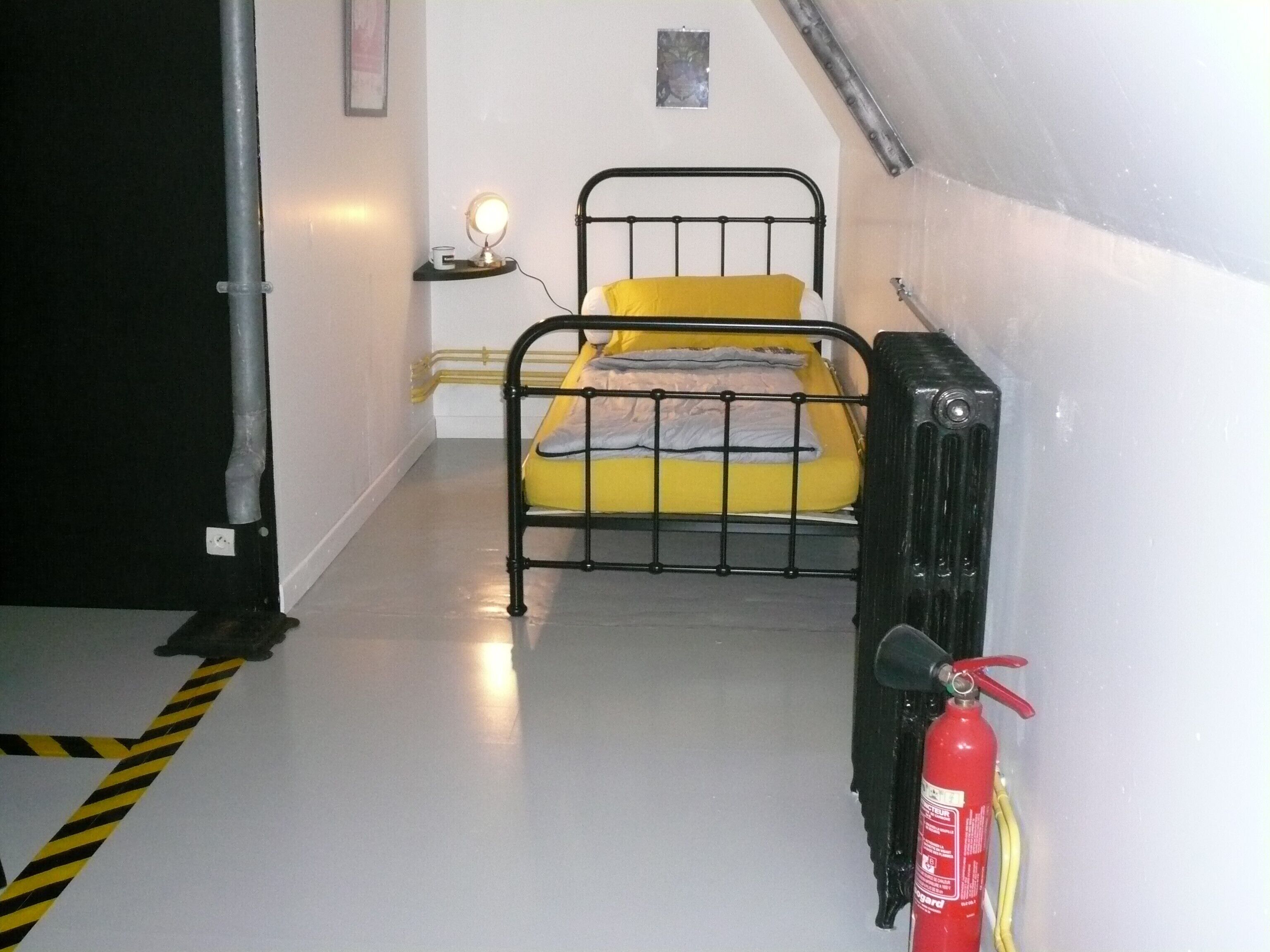 A small, single-bed room with a yellow bedspread, a bedside table with a lamp, a red fire extinguisher, and a radiator.