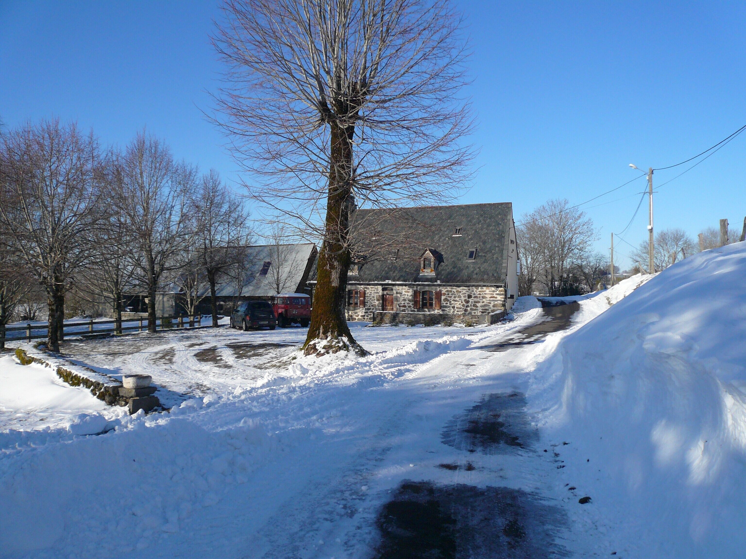 A snow-covered rural road with a stone building, bare trees, and parked cars.