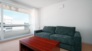 A living room with a sofa, a wooden coffee table, a window with a view of buildings, and a white wall. - Small studio for 2 people, close to amenities (Agde)
