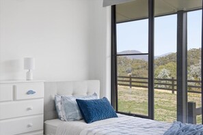 A bedroom with a bed, a nightstand, a lamp, and a large window with a view of a fenced area and trees. - Eildon Valley Views - in the heart of the scenic Eildon Valley (Eildon)