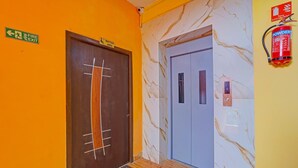 A hallway with a fire exit sign, a waiting area sign, a fire extinguisher, and a door with a number plate. - Deluxe tad Homes (Sriperumbudur)