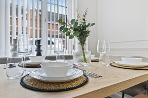 Une table à manger dressée avec des assiettes, des bols, des couverts et des verres à vin, et un vase rempli de verdure en arrière-plan.