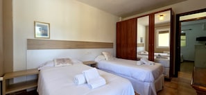 A hotel room with two beds, a wooden headboard, a bedside table, a mirror, and a door leading to another room.