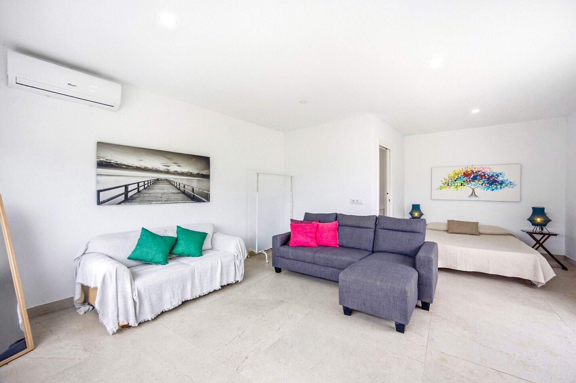 A spacious room with a white couch, a grey sofa, and a bed. There are two paintings on the walls, a whiteboard, and a ceiling-mounted air conditioning unit.
