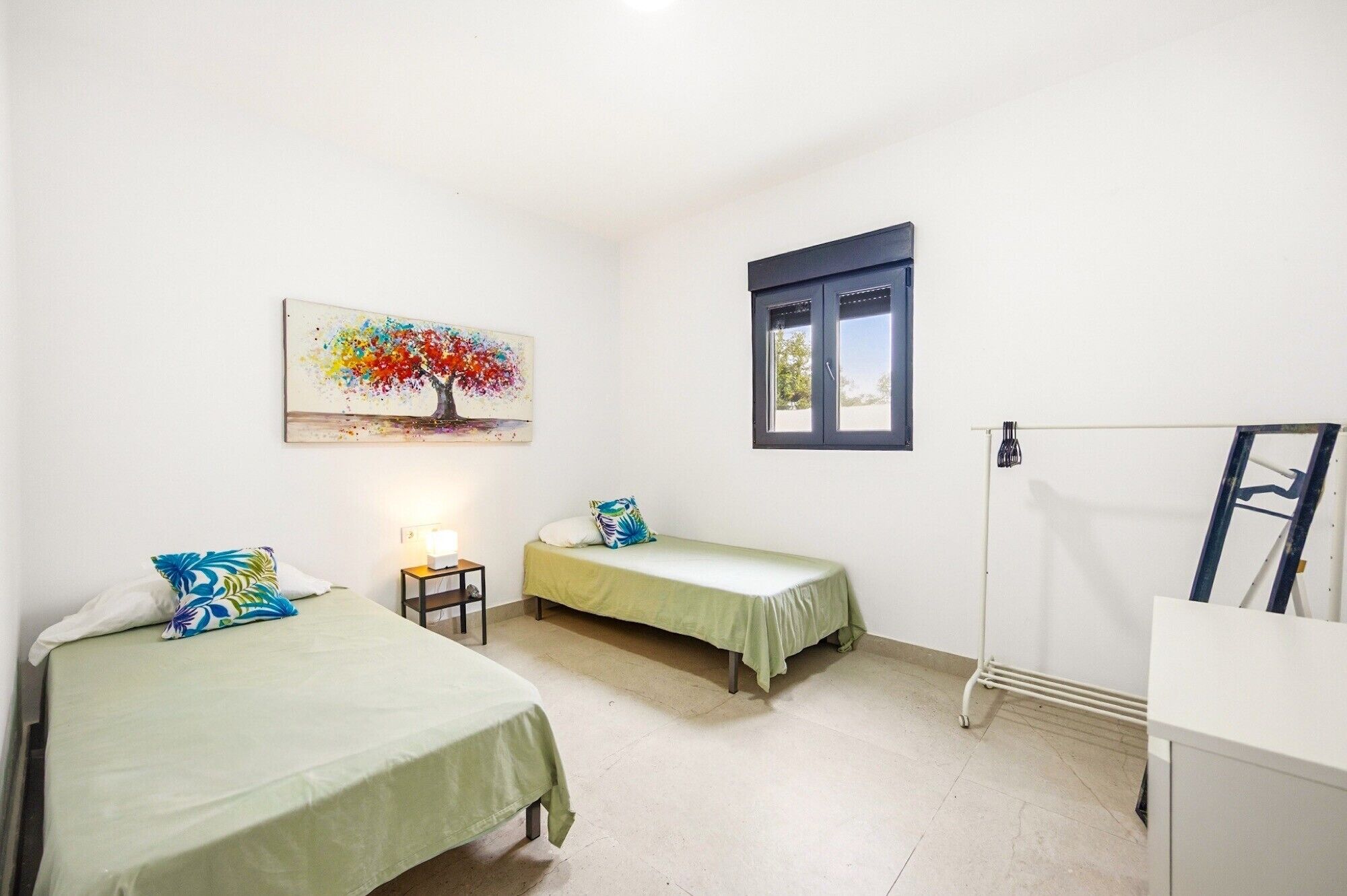A bedroom with two beds, a nightstand, a lamp, a window, and a painting of a tree.