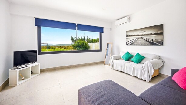 A modern living room with a large window, a flat-screen TV, a sofa, an ottoman, and a painting on the wall.