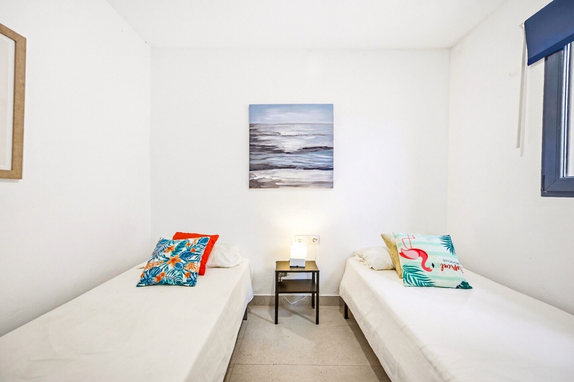 A small, clean room with two single beds, a nightstand, a wall-mounted painting, and a window with a blue frame.
