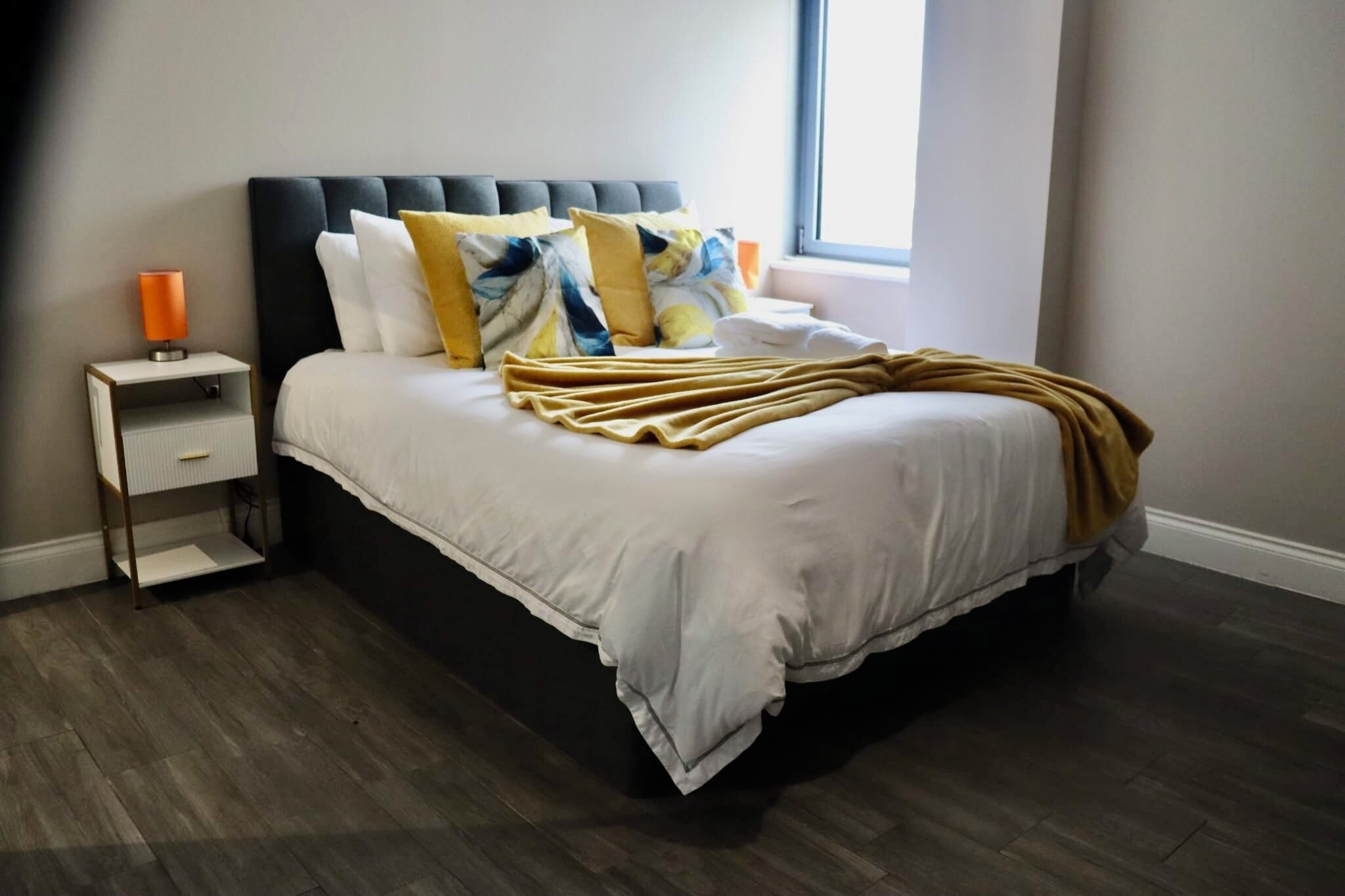 A neatly made bed with a headboard, a bedside table with a lamp, and a window providing natural light.