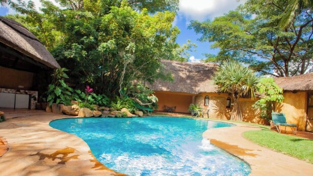 A swimming pool surrounded by a stone patio, lush greenery, and a thatched-roof building. - Green Oasis Lodges (Harare)
