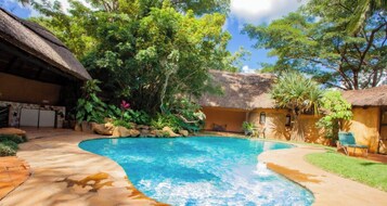 Una piscina rodeada por un patio de piedra, abundante vegetación y un edificio con techo de paja.