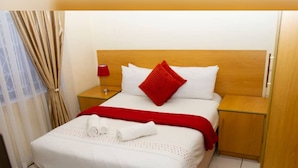 A hotel room with a bed, a nightstand, a lamp, a window with curtains, and a door. - Green Oasis Lodges (Harare)