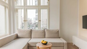 A modern living room with a beige sofa, a small wooden table with a bowl of fruit, and a TV on the wall.
