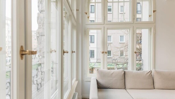 A room with a large window, a beige sofa, and a view of buildings outside.